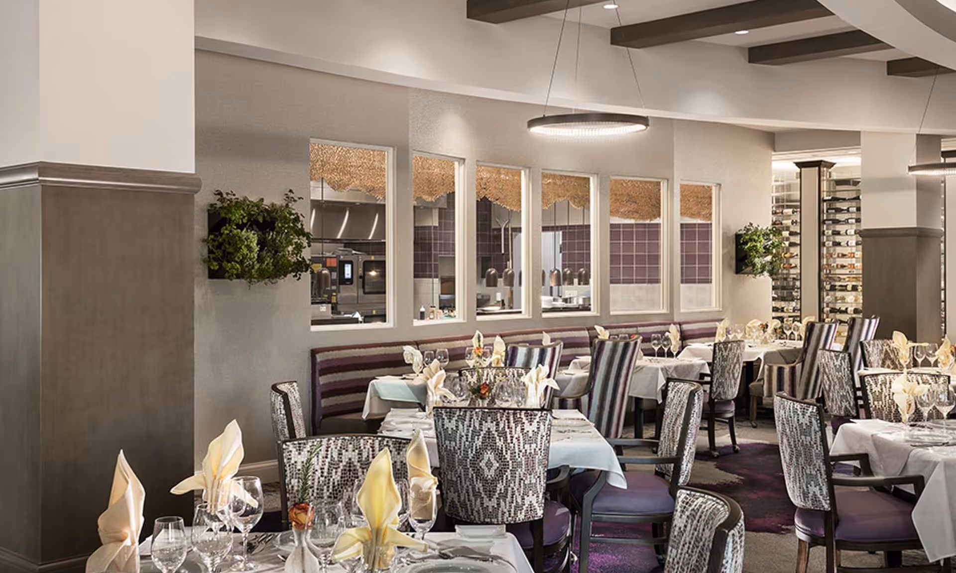 Elegant dining room with tables set with white tablecloths, folded napkins, glassware, and floral centerpieces. The room features patterned chairs, a wine rack, and windows looking into a kitchen area. Modern circular pendant lights hang from the ceiling.