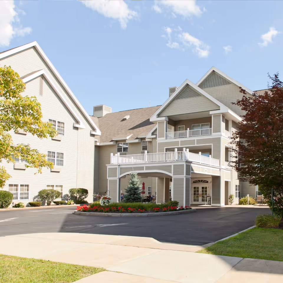 Exterior view of Parkrose Estates, a multi-story senior living facility with beige siding, white trim, and a covered entrance. The building is surrounded by landscaped greenery, including trees, bushes, and a flower bed with red flowers. The sky is clear with a few clouds.