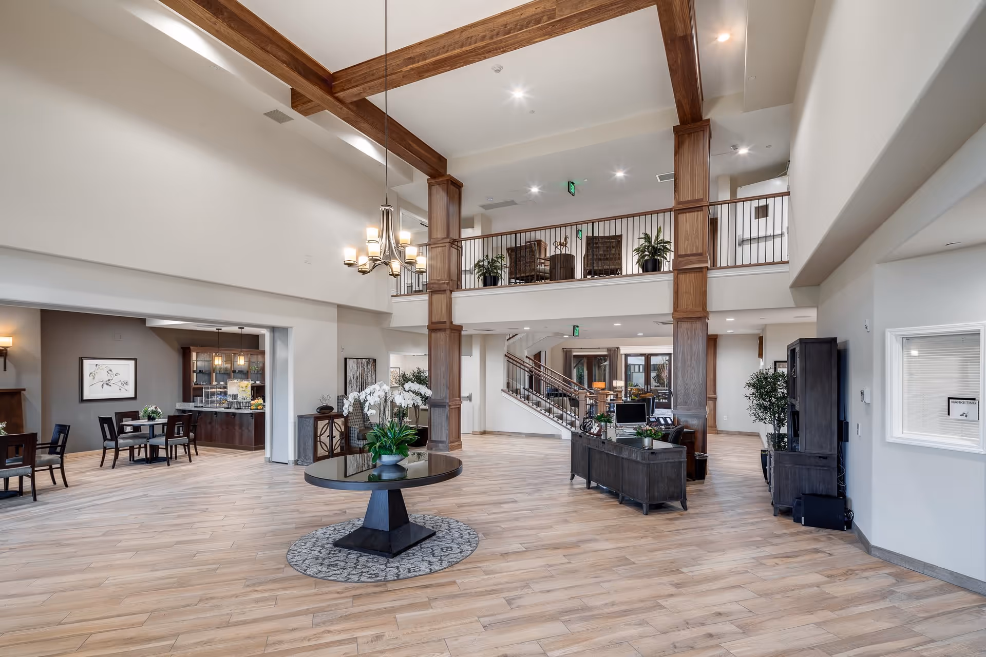 Spacious and well-lit interior of a senior living facility with high ceilings and wooden beams. The room features a round table with a flower arrangement in the center on a patterned rug, a chandelier hanging from the ceiling, and a reception desk with computer monitors. There is a staircase leading to an upper balcony area with seating and plants. To the left, there is a dining area with tables and chairs, and to the right, a window labeled 'Marketing'. The flooring is light wood, and the walls are painted white.