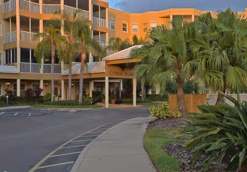 Entrance of a multi-story senior living building labeled Cypress Palms with palm trees and a curved driveway.