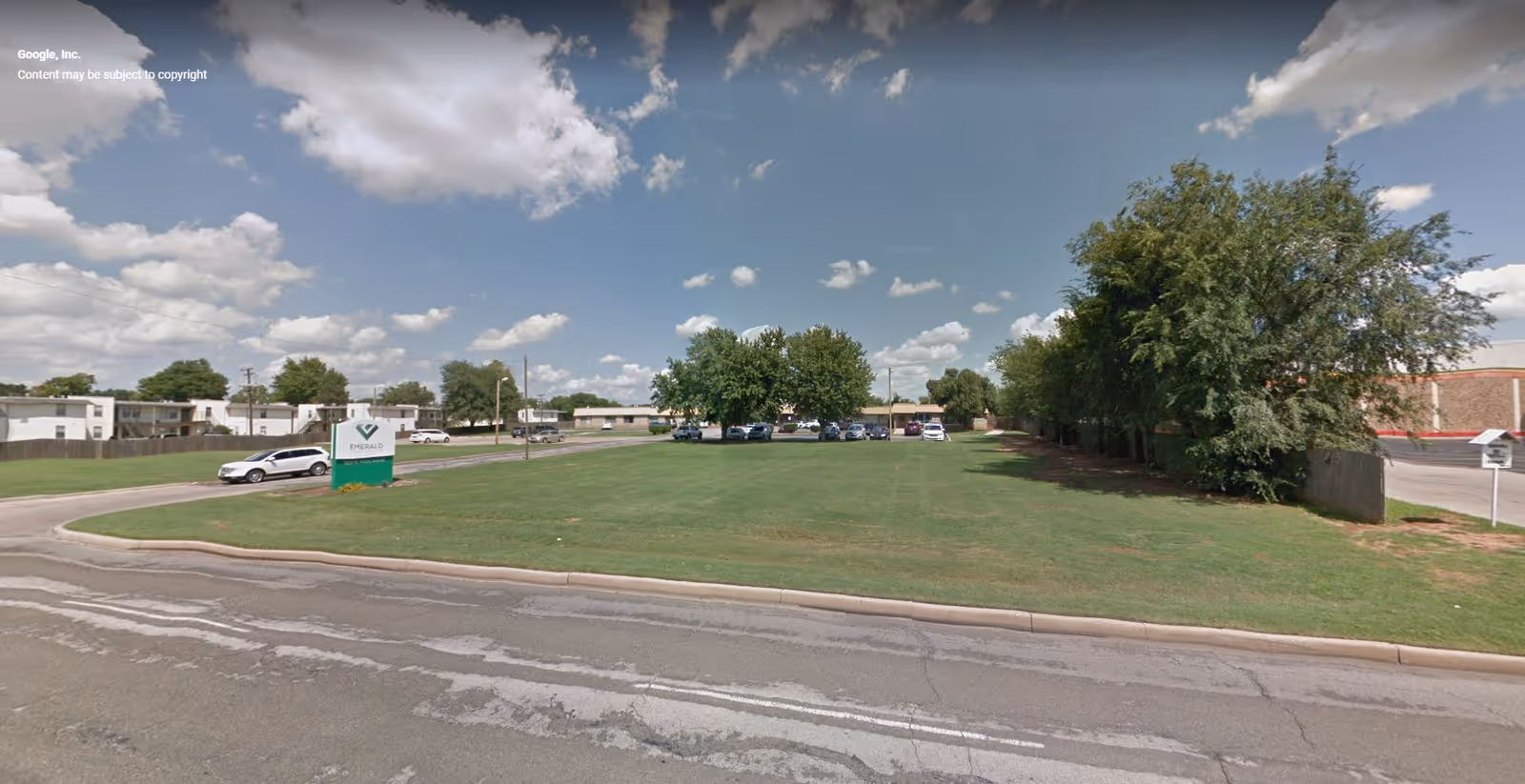 Exterior view of Emerald Care Center Southwest showing a large grassy area with trees on the right side, a parking lot with several cars in the background, and a sign with the facility's name near the street under a partly cloudy sky.