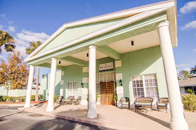 Front entrance of a light-green building with tall white columns, a covered portico and outdoor seating.