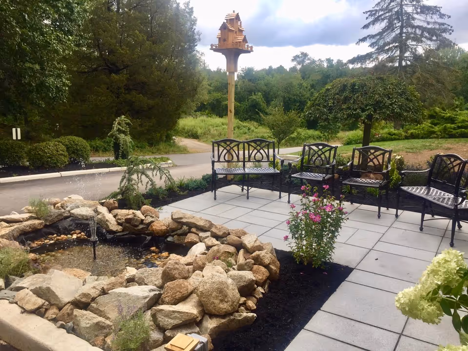 Outdoor patio with black metal chairs, a small rock fountain surrounded by rocks and flowerbeds, and a tall birdhouse in a landscaped garden.