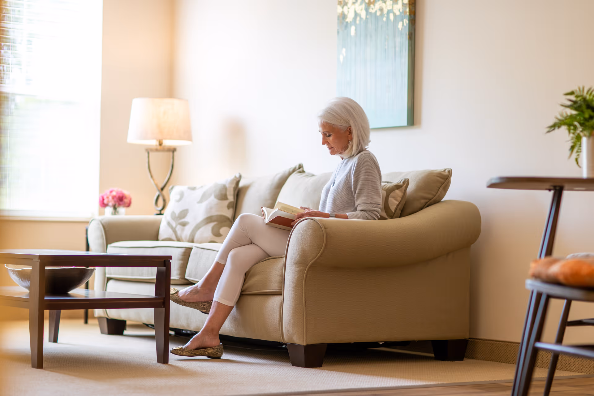 An elderly woman with white hair sitting on a beige sofa in a well-lit living room, reading a book. The room has a wooden coffee table, a floor lamp, a painting on the wall, and a small table with a plant and some items on it.