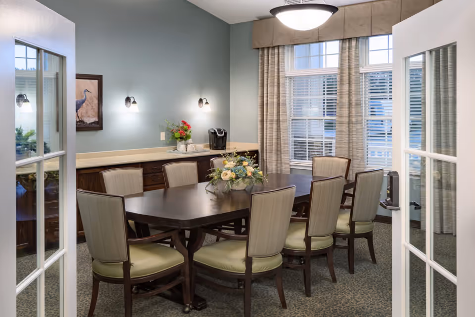 A well-lit dining room with a large dark wooden table surrounded by eight cushioned chairs. The room has light green walls, a sideboard with a coffee maker and a flower arrangement, two wall sconces, a framed picture of a bird, and large windows with beige curtains allowing natural light to enter. The view is through open French doors.