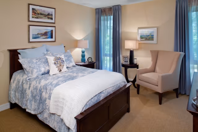 A cozy bedroom with a wooden bed frame, neatly made bed with blue and white floral bedding and multiple pillows. There are two framed landscape paintings on the wall above the bed. A nightstand with a blue lamp and an alarm clock is beside the bed. Next to a window with blue curtains, there is a beige upholstered armchair and a small round table with a lamp and a framed photo. The room has beige walls and carpeted floor.