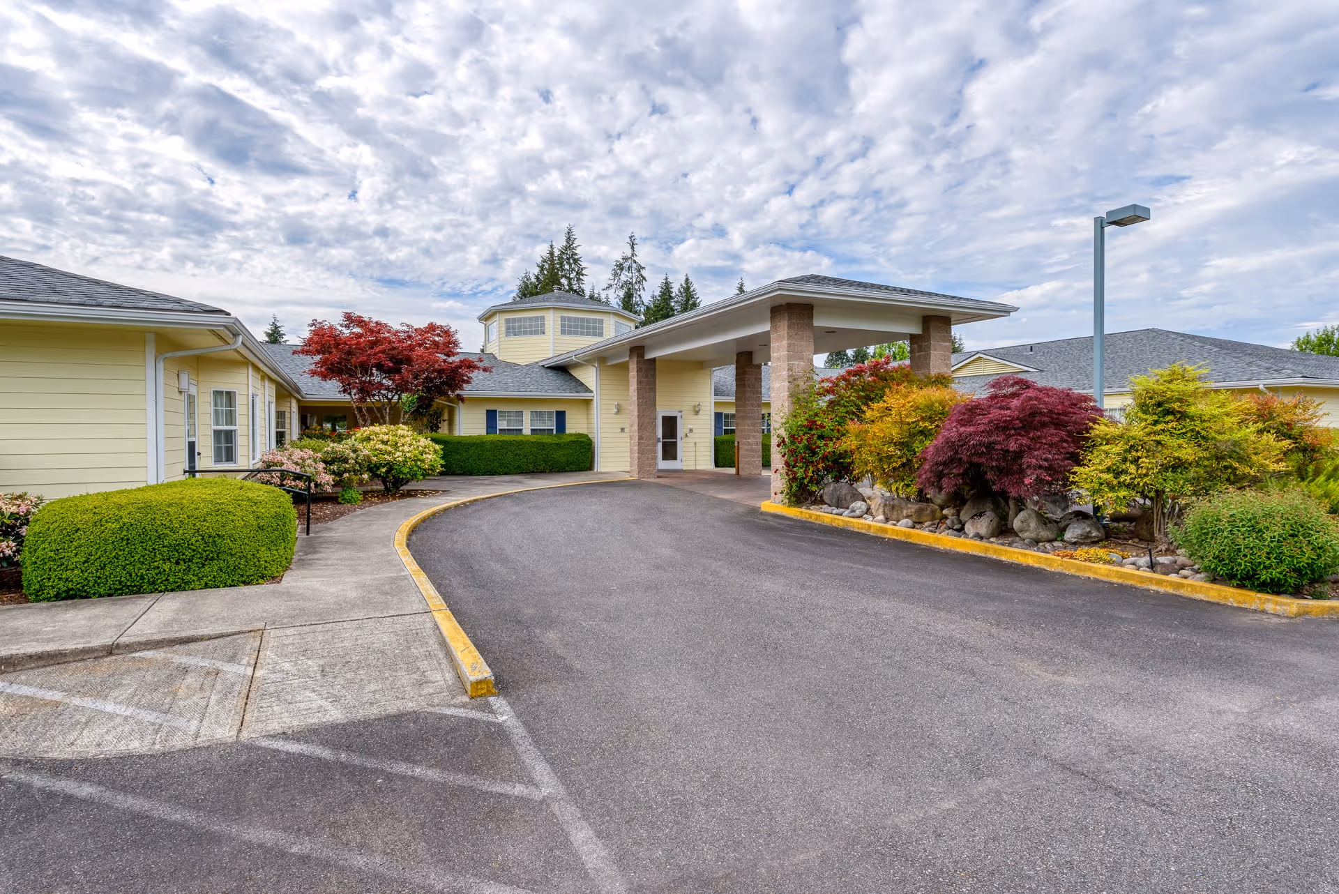 Entrance of Easthaven Villa Assisted Living & Memory Care facility with a covered drop-off area, surrounded by well-maintained landscaping including bushes and colorful trees under a partly cloudy sky.