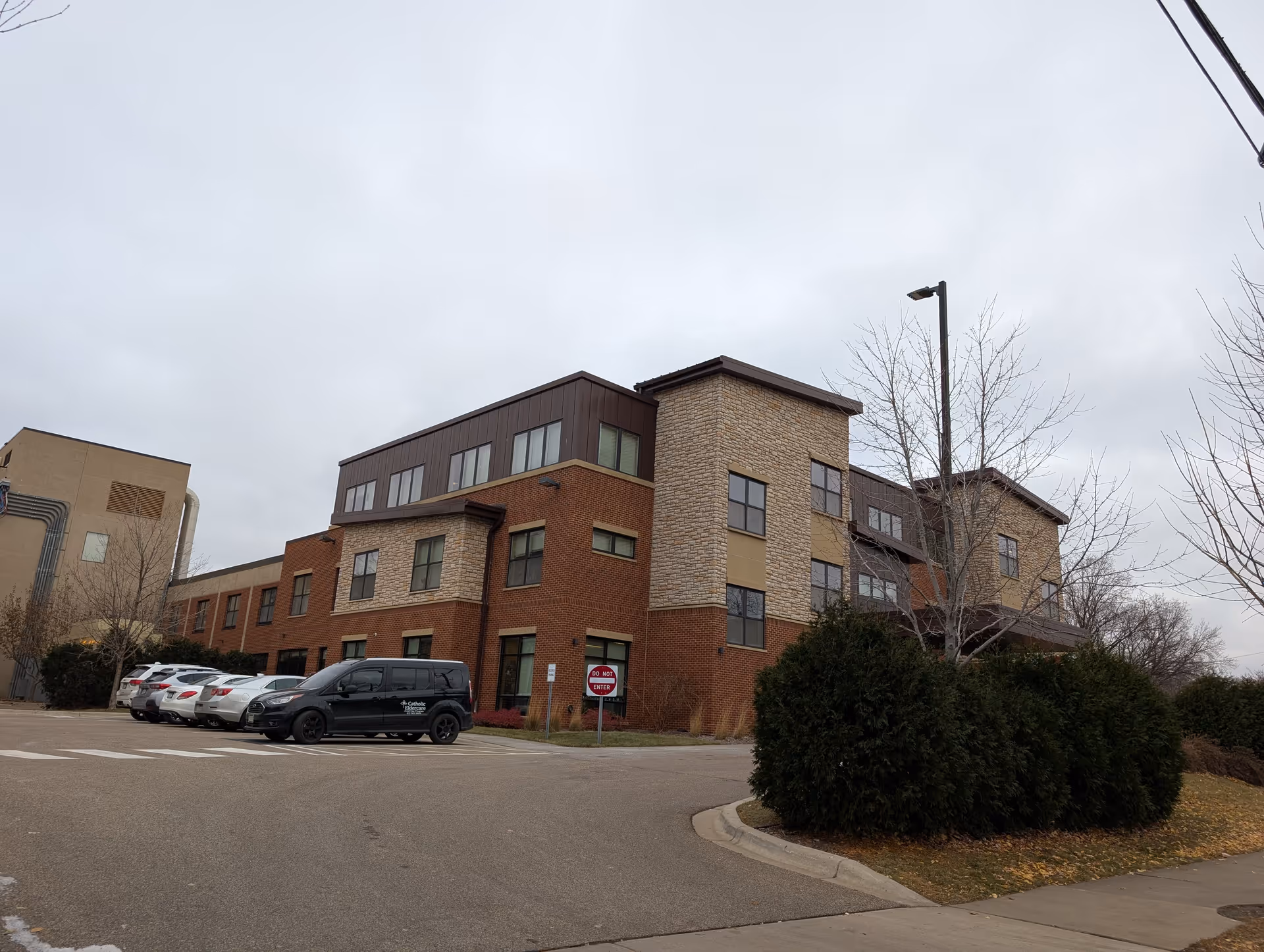 Three-story brick and stone senior living building with parked cars and shrubs by the driveway under an overcast sky.