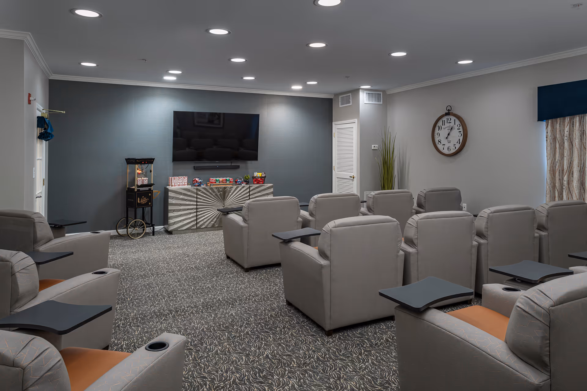 A small theater-style room with multiple rows of gray reclining chairs, each equipped with a small black tray table. At the front of the room is a large flat-screen TV mounted on a gray accent wall, with a popcorn machine and a cabinet below it holding various snacks. The room has recessed ceiling lights, a large wall clock, and a window with patterned curtains and a dark blue valance.