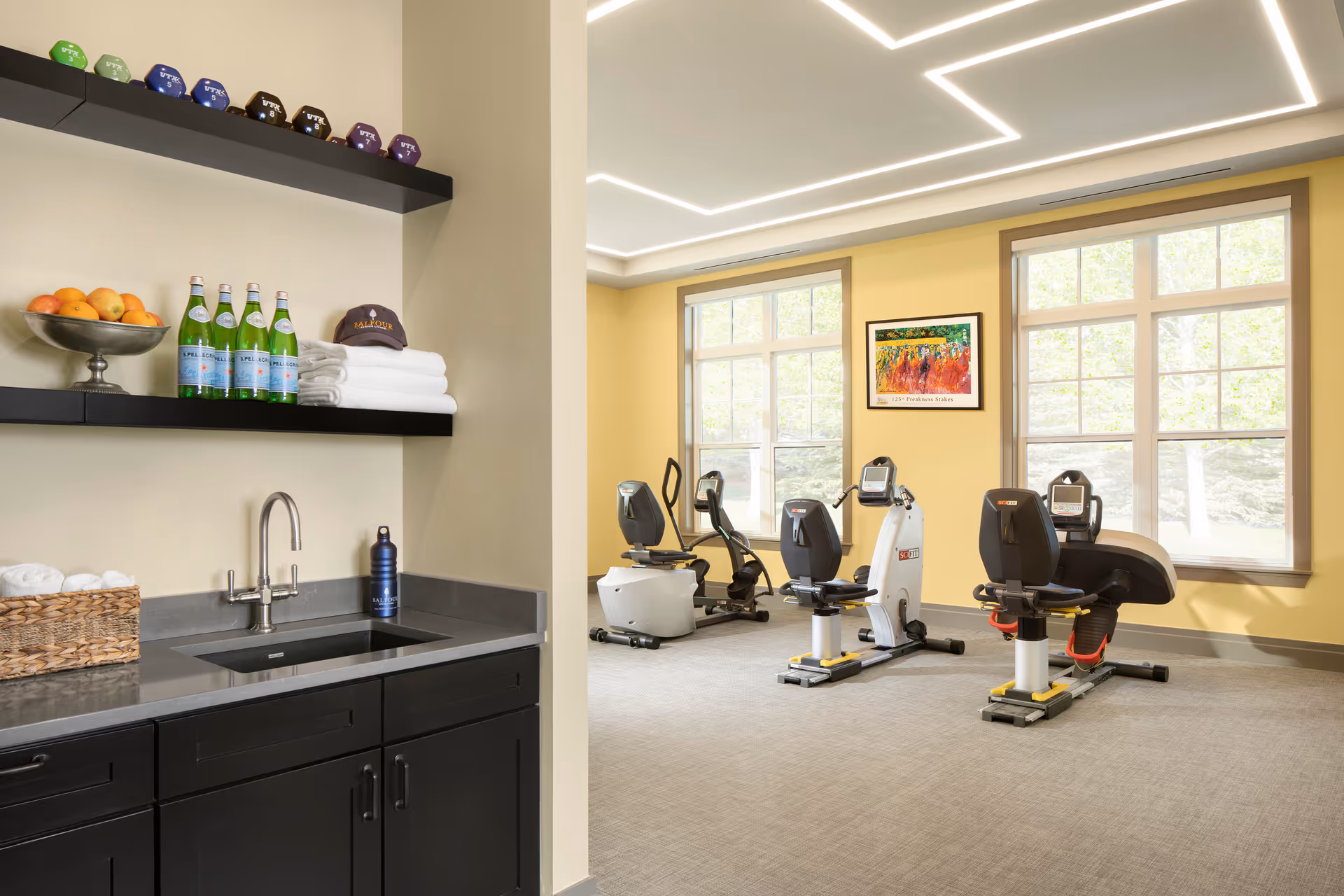 A bright exercise room with three stationary recumbent bikes positioned near large windows letting in natural light. The walls are painted yellow, and there is a colorful framed painting on the wall. In the foreground, there is a black countertop with a sink, shelves holding bottles of sparkling water, a bowl of fruit, folded towels, and a cap with the name 'Balfour' on it.