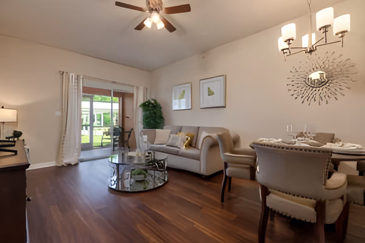 A cozy living and dining area with a beige sofa, a glass coffee table, and a dining table with upholstered chairs. The room has wooden flooring, a ceiling fan with lights, and a chandelier above the dining table. Large sliding glass doors lead to an outdoor area with greenery visible outside.