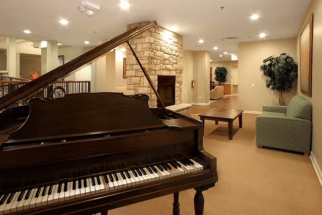Interior view of a supportive living facility featuring a grand piano in the foreground, a stone fireplace in the background, beige walls, recessed lighting, a green upholstered chair, a wooden coffee table, and potted plants along the hallway.