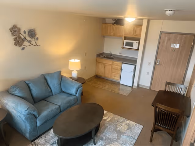 A small living area with a blue loveseat, a round coffee table on a patterned rug, and a side table with a lamp. In the background, there is a kitchenette with wooden cabinets, a microwave, a small refrigerator, and a sink. To the right, there is a wooden dining table with two chairs and a closed wooden door.