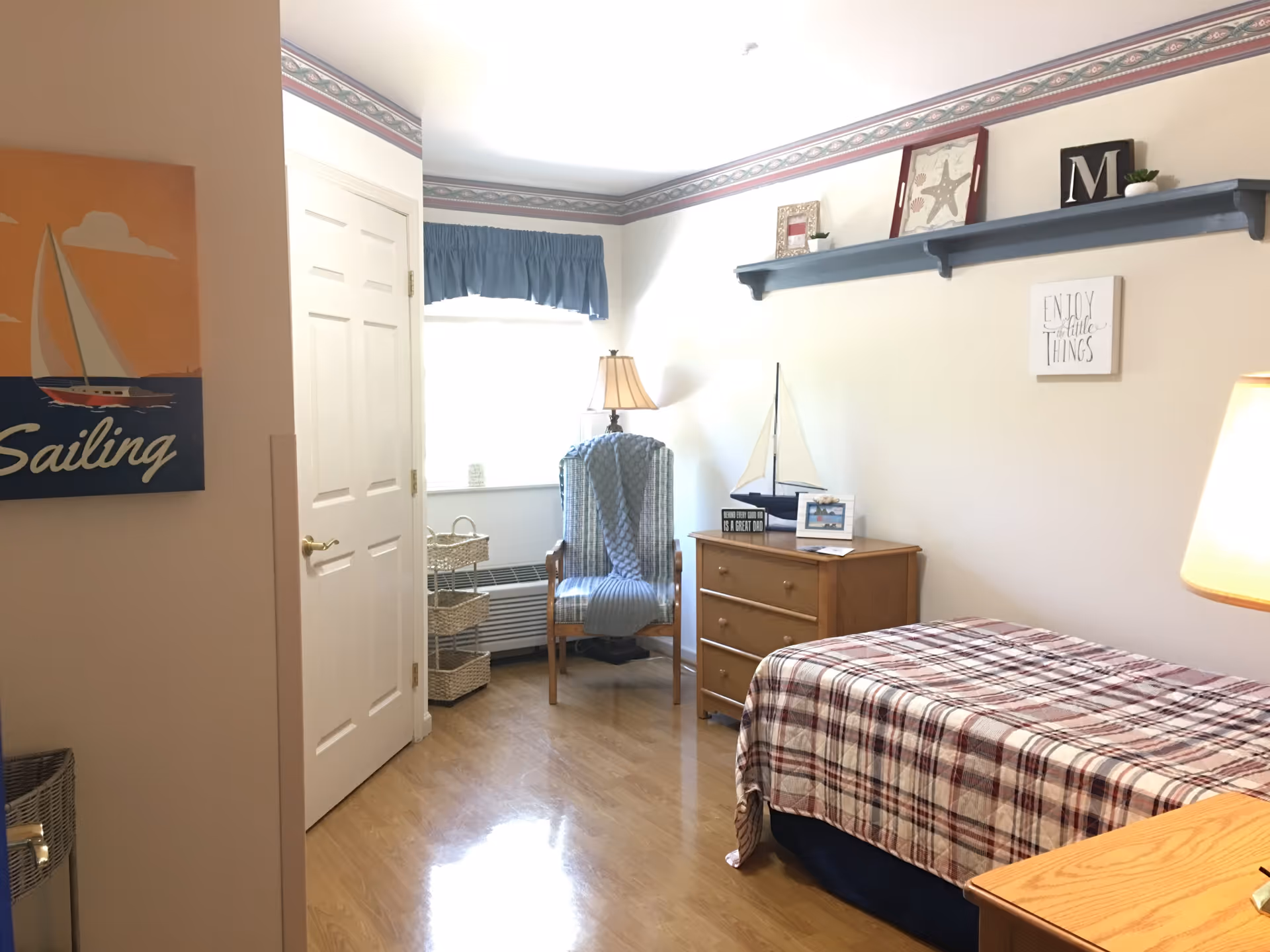 A tidy single-occupancy bedroom with a twin bed covered in a plaid blanket, wooden dresser and armchair by a window with nautical decor.