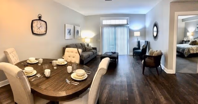 A cozy living and dining area in a retirement community apartment. The dining area features a round wooden table set for four with plates, bowls, cups, and cutlery. Adjacent to the dining area is a living room with a gray sofa, a cushioned chair, a coffee table, and a floor lamp. The room has wooden flooring and light-colored walls with framed artwork and a clock. Sliding glass doors with vertical blinds lead to an outdoor space. A bedroom is partially visible through an open doorway.