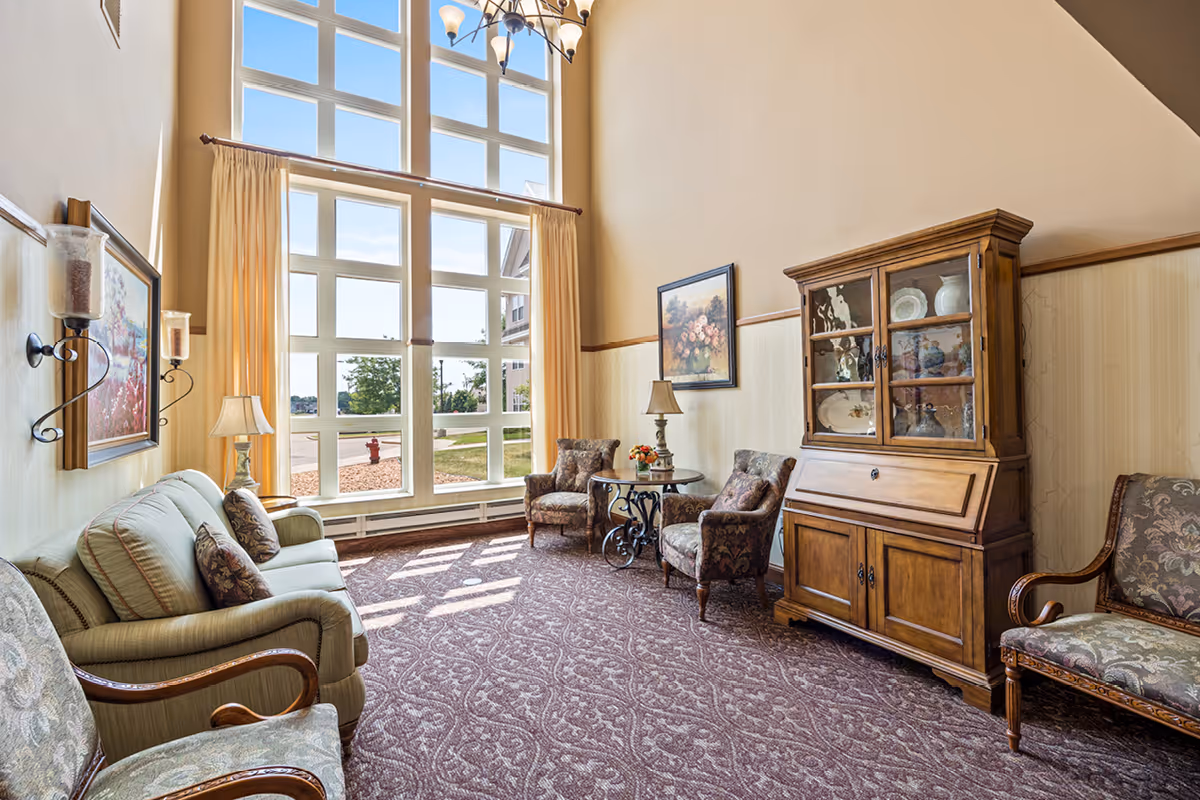 A bright and cozy living room with large floor-to-ceiling windows letting in natural light. The room features a patterned carpet, a green sofa with decorative pillows, two upholstered armchairs around a small round table with a lamp and flower arrangement, a wooden cabinet with glass doors displaying dishes, and framed artwork on the walls.