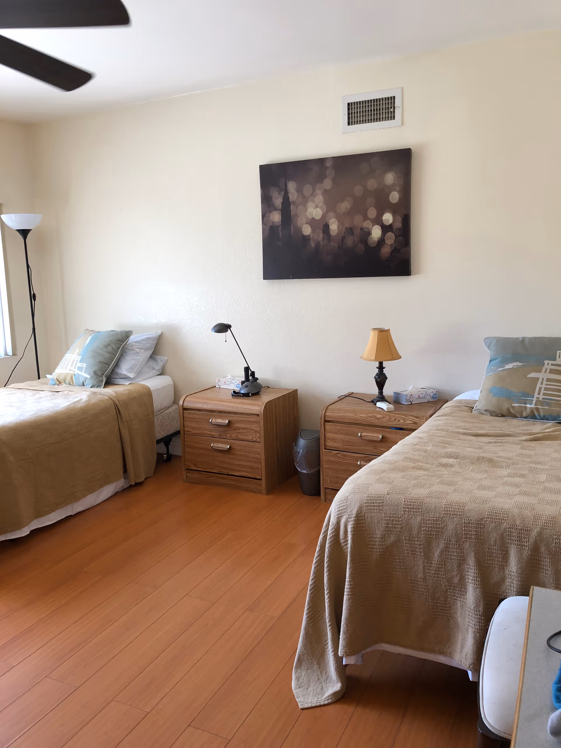 A bedroom with two single beds, each with beige bedspreads and decorative pillows. Between the beds are two wooden nightstands with lamps and a box of tissues. A floor lamp stands near the window on the left, and a framed artwork hangs on the wall above the nightstands. The floor is wooden, and the walls are light-colored.