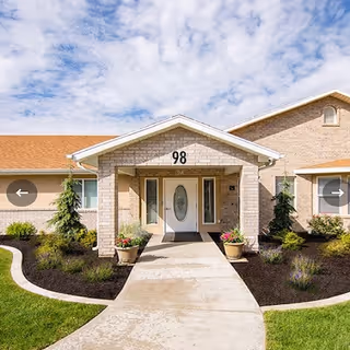 Front entrance of a brick senior living facility with a curved walkway and landscaped garden beds.