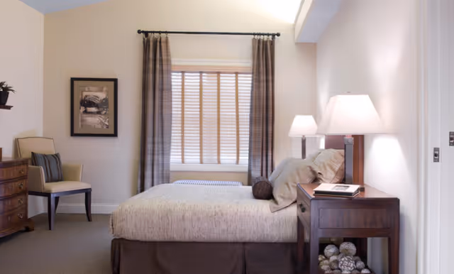 Bright, neatly arranged bedroom with a bed, wooden nightstand and lamps, a chair and window with curtains.