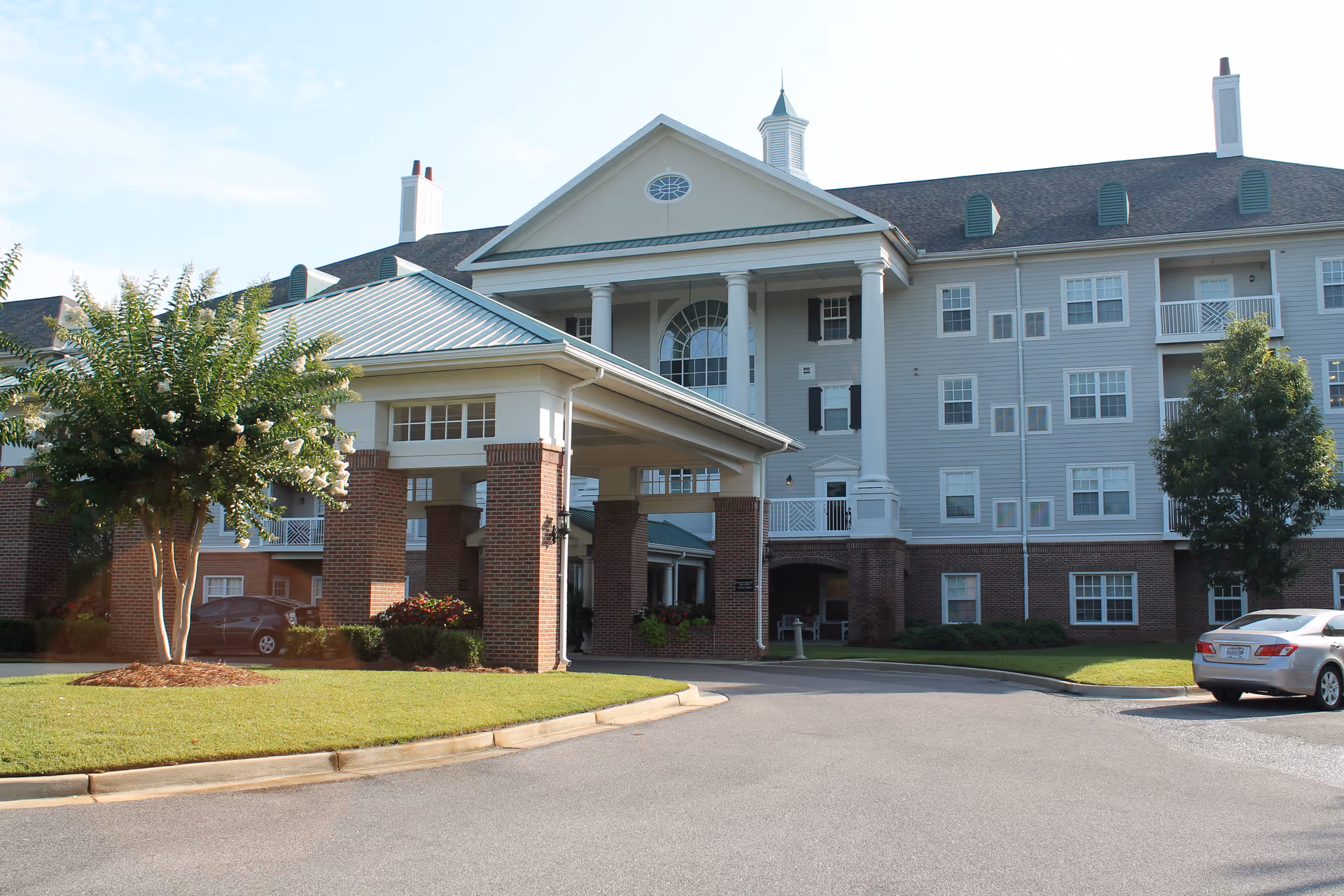 Exterior view of a large senior living facility building with a covered entrance supported by brick columns. The building has multiple windows, a pitched roof with chimneys, and a small cupola on top. There are trees and shrubs around the entrance, and a car is parked on the driveway.