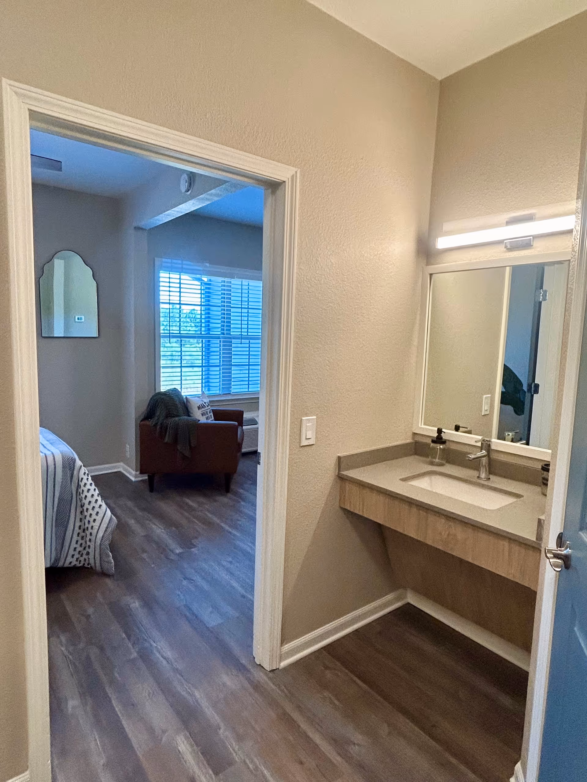 View of a small vanity area with a sink and mirror on the right, leading into a bedroom with a bed, a brown armchair, and a window with blinds letting in natural light.