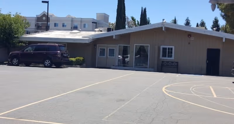 Exterior view of a single-story building with beige siding and a slanted roof, adjacent to a parking lot with marked spaces and a basketball court. A dark SUV is parked near the building, and there are tall trees and a multi-story building in the background.