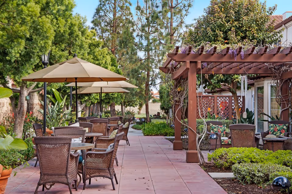 Outdoor patio area with multiple tables and wicker chairs under large beige umbrellas. There is a wooden pergola with climbing plants and colorful cushions on chairs. The area is surrounded by green trees, bushes, and plants, creating a peaceful garden setting.