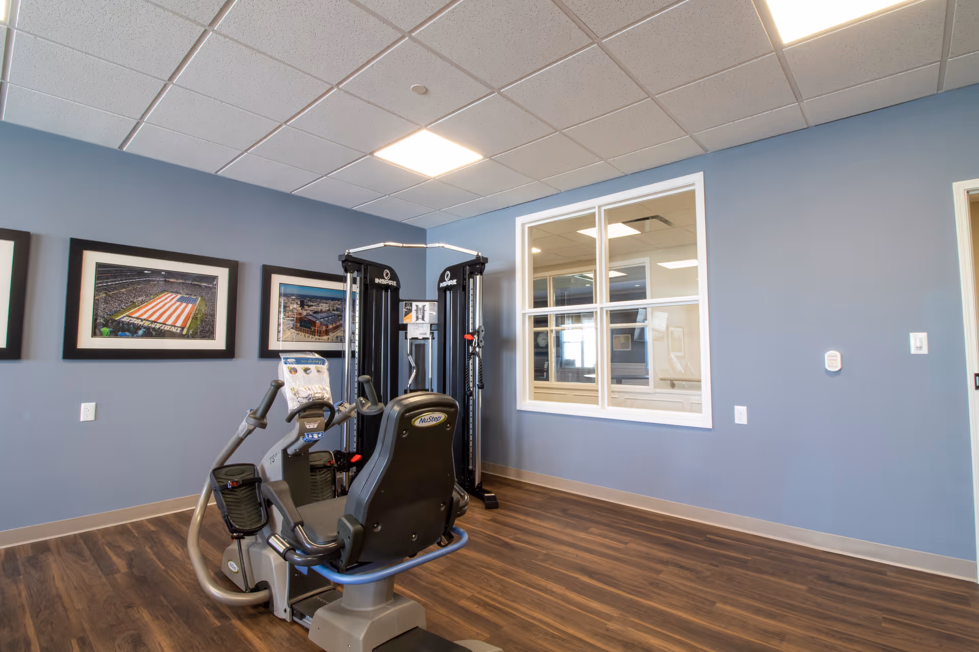 A small fitness room with exercise equipment including a recumbent bike and a multi-function weight machine. The room has blue walls, wood flooring, a window looking into another room, and framed pictures on the wall.