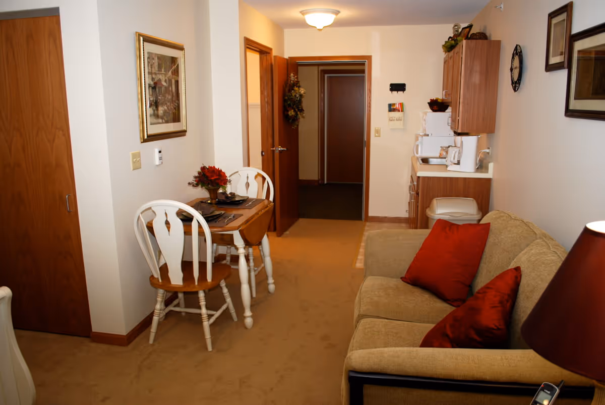 Interior view of a senior living facility room at Heritage Woods of McHenry featuring a small dining area with a wooden table and two chairs, a beige couch with red pillows, a kitchenette with cabinets, a microwave, and a coffee maker, and a hallway with open doors leading to other rooms.