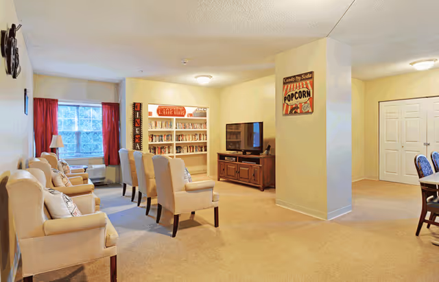 Common area with rows of armchairs facing a television, bookshelves, and a dining area in a senior living facility.