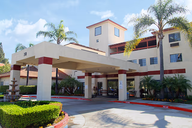 Exterior view of a senior living facility with a covered entrance supported by columns. The building is beige with red tile accents and surrounded by palm trees and landscaped greenery under a blue sky.