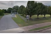 Paved driveway with grassy median, trees, and a sign at the entrance to a senior facility.