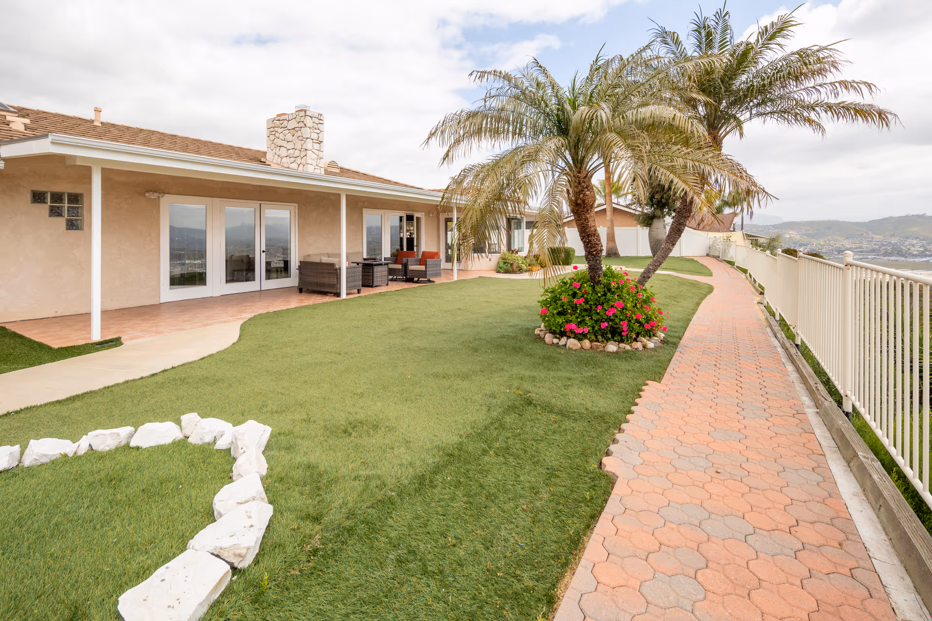 Outdoor view of a senior living facility with a paved walkway alongside a green lawn featuring palm trees and flower beds. The building has a covered patio area with outdoor seating and a stone chimney, overlooking a scenic landscape with hills in the background.