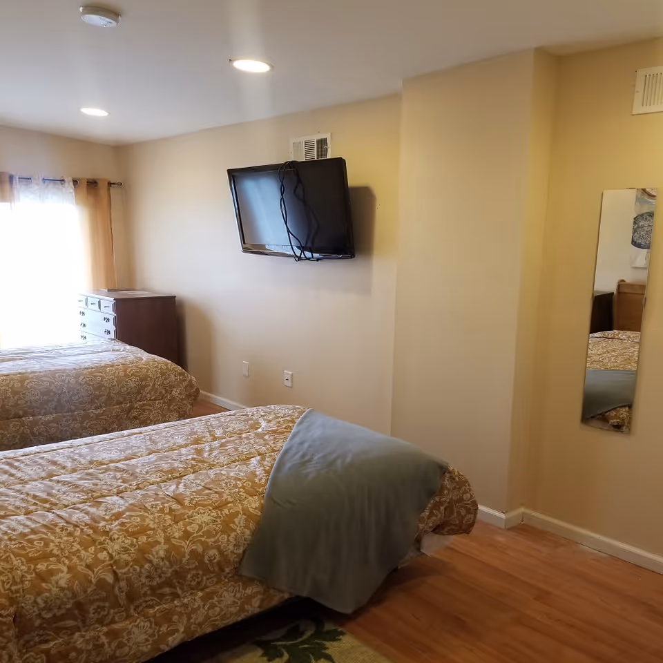 A bedroom with two beds, a wall-mounted TV, a dresser by a curtained window, and a full-length mirror.