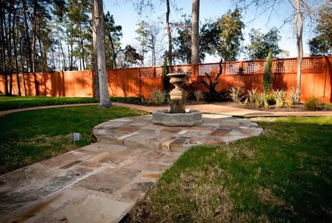A peaceful outdoor garden area with a stone pathway leading to a circular stone platform featuring a decorative water fountain. The garden is surrounded by a wooden fence and trees, with green grass and some plants along the fence.