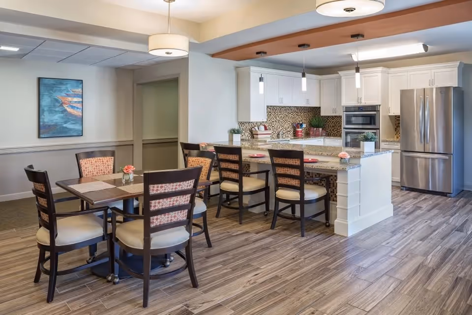 A modern kitchen and dining area in a senior living facility with a wooden dining table and six chairs, a kitchen island with four chairs, white cabinets, stainless steel refrigerator and oven, pendant lights, and a decorative painting on the wall.