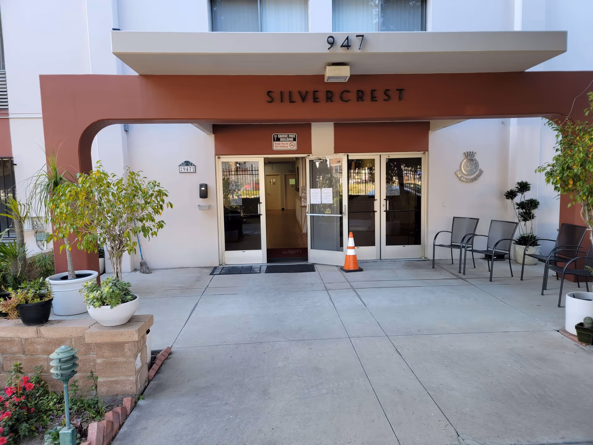 Entrance to The Salvation Army LA Silvercrest Senior Housing Residence with double glass doors, a brown overhang with the number 947 and the word SILVERCREST above the doors. There are potted plants on both sides of the entrance and several chairs on the right side. A traffic cone is placed in front of one of the doors.