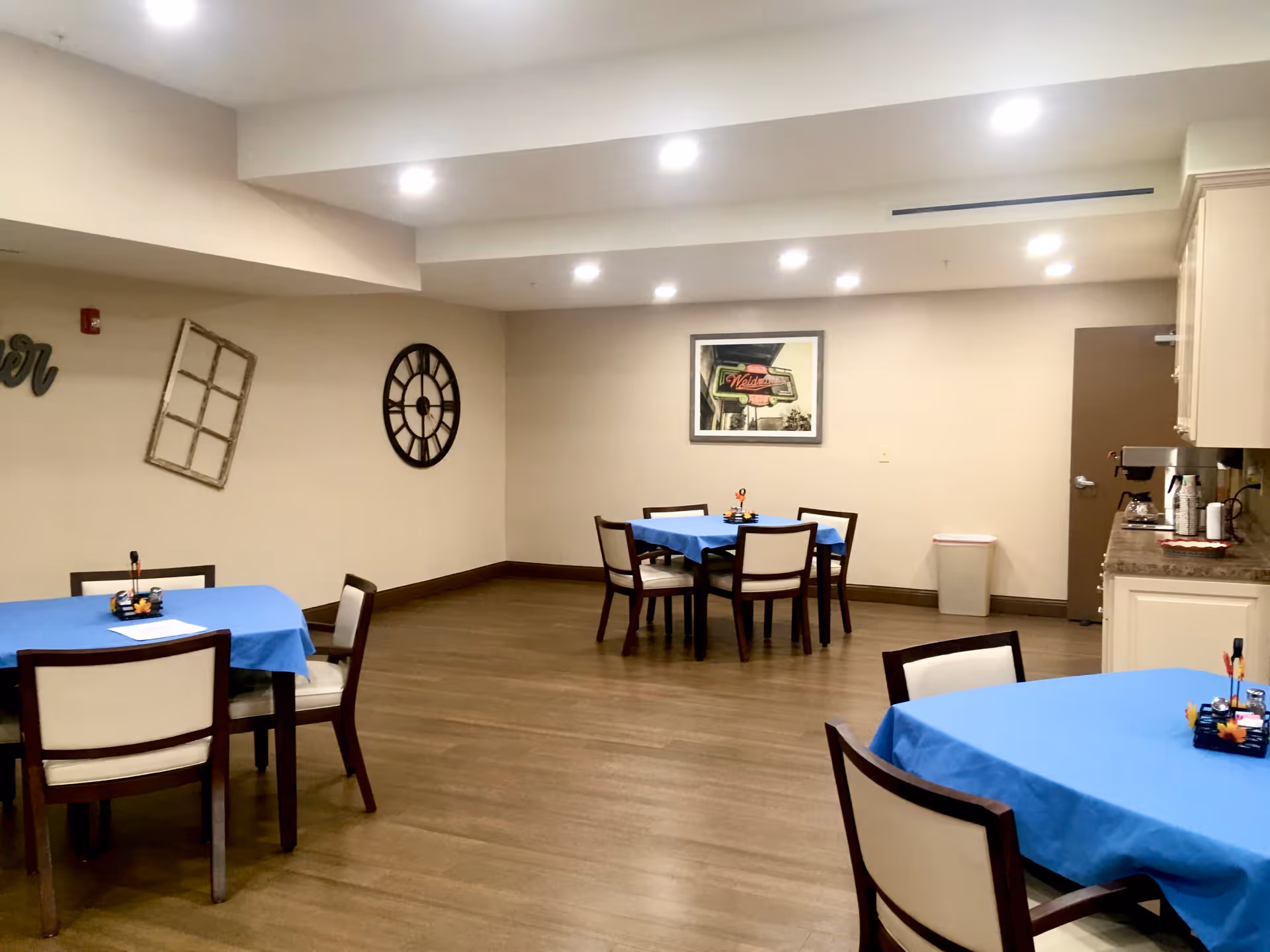 Community dining room with several small tables covered in blue tablecloths, chairs, wall decor, and a kitchenette area.