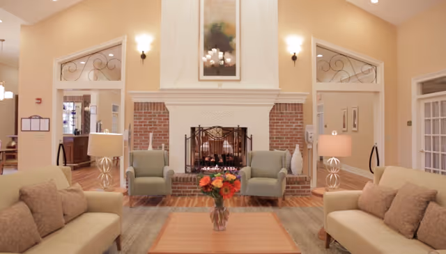 A cozy living room area with two beige sofas and two green armchairs arranged around a wooden coffee table with a vase of colorful flowers. The room features a brick fireplace with a white mantel and a painting above it. Two table lamps with beige shades are placed on side tables next to the sofas. The walls are painted a warm beige color, and the floor is wooden with a large area rug.