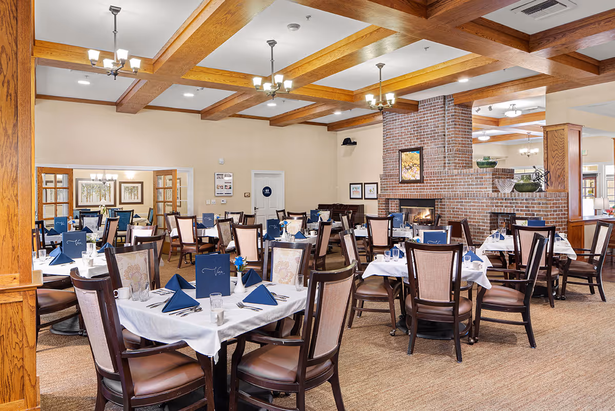 A spacious dining room with multiple tables covered in white tablecloths, each set with blue napkins, menus, glasses, and silverware. The room features wooden ceiling beams, chandeliers, a brick fireplace with a painting above it, and beige walls. The chairs have dark wooden frames with cushioned seats and backs. There are framed pictures on the walls and a carpeted floor.