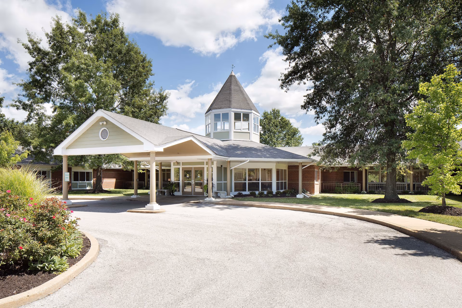 Front entrance of a single-story senior care building with a covered porte-cochère, a small tower, trees, and landscaped grounds under a blue sky.