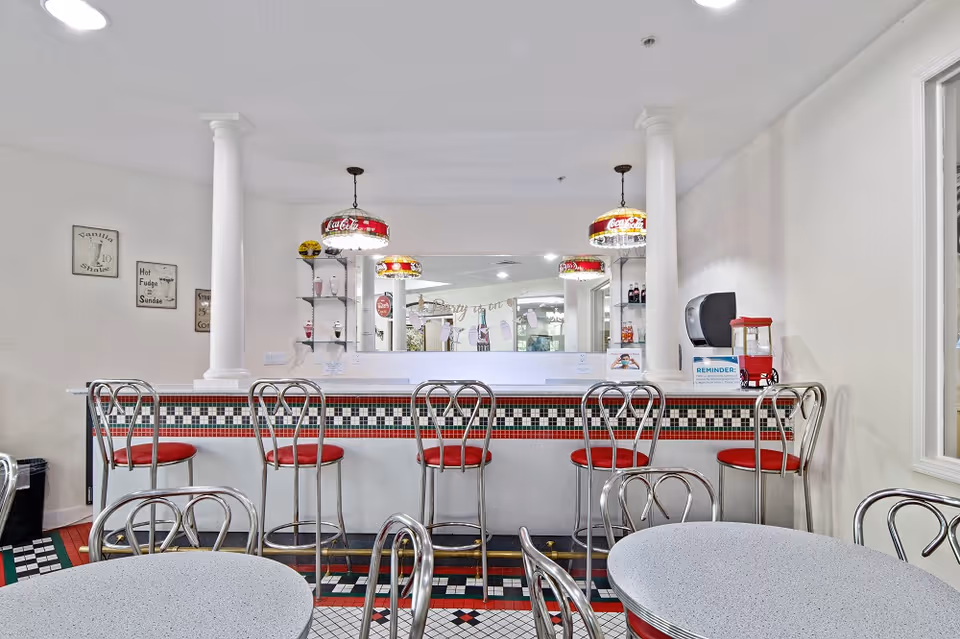 Retro-style indoor diner counter with red cushioned barstools, Coca-Cola pendant lamps, chrome chairs and small round tables.