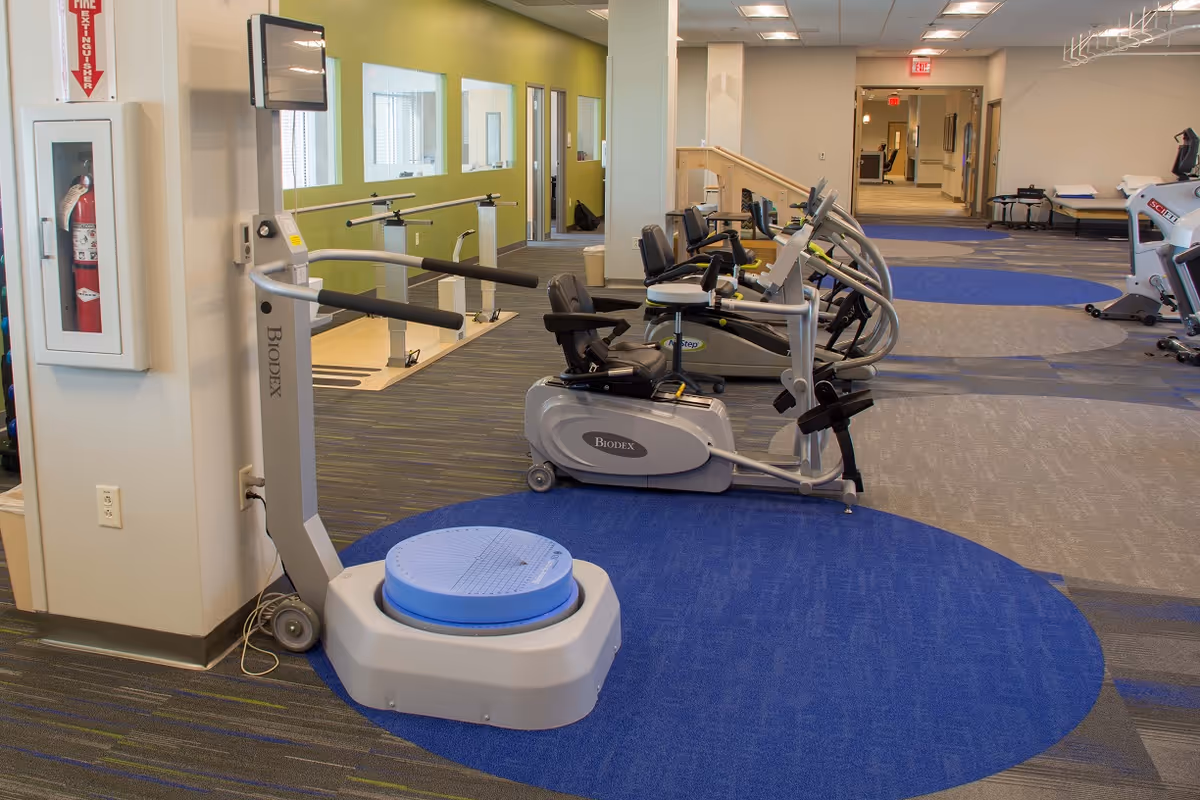 Interior view of a fitness and rehabilitation room with exercise equipment including stationary bikes and a Biodex balance testing machine. The room has a blue and gray carpet with circular patterns, a green accent wall with windows, and a fire extinguisher mounted on a white pillar.