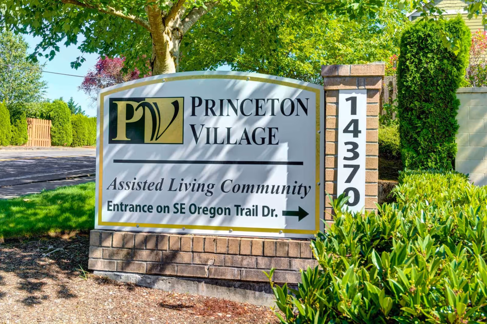 Entrance sign for Princeton Village assisted living community next to a brick address pillar and landscaped greenery.