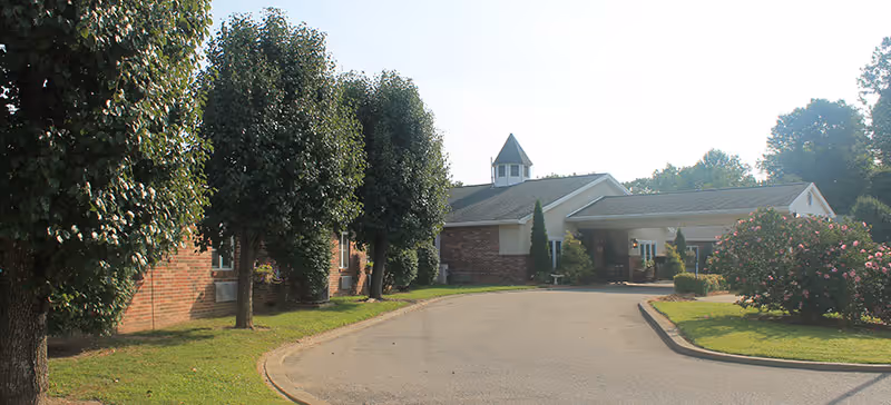 Front entrance of a brick senior living building with a covered driveway and landscaped trees and shrubs.