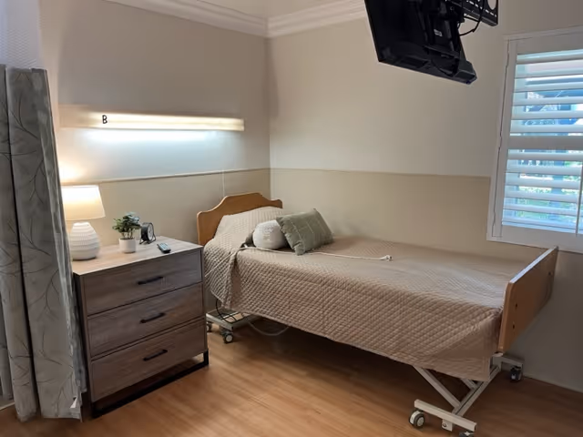 A simple, clean patient room with a single hospital bed covered in a beige quilted blanket and two pillows. Next to the bed is a wooden nightstand with three drawers, a table lamp, a small plant, and a clock. A wall-mounted TV is positioned above the bed, and a window with white shutters lets in natural light. The floor is wooden, and the walls are painted in neutral tones.