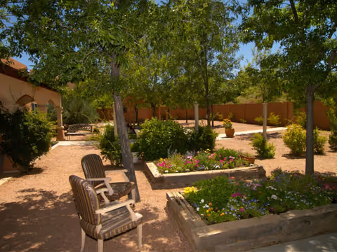 Outdoor garden area with raised flower beds filled with colorful flowers, several trees providing shade, two striped cushioned chairs, and a gravel pathway. A brown fence and part of a building are visible in the background.