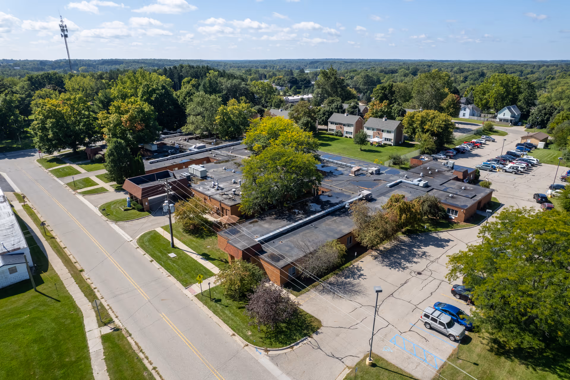 Aerial view of Intersect Healthcare of Belding, showing a single-story brick building with a flat roof surrounded by trees, parking lots with cars, and nearby residential houses under a clear blue sky.