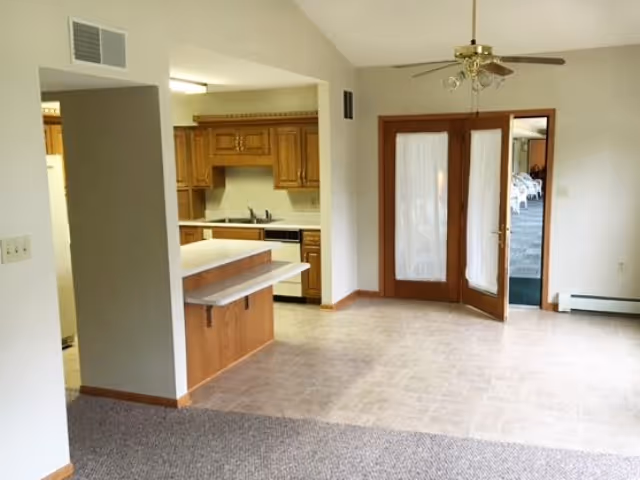 Interior view of a senior living facility showing a kitchen area with wooden cabinets and a countertop, adjacent to an open space with tiled flooring and carpeted area. There are double glass doors with white curtains leading to another room, and a ceiling fan with lights is mounted above.