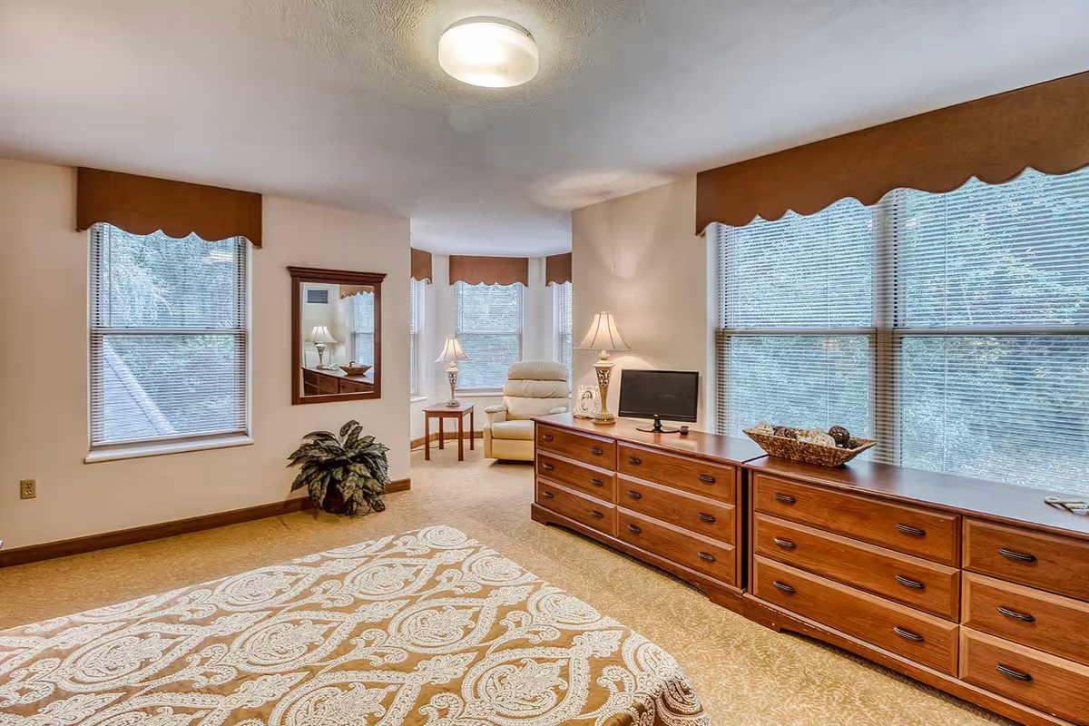 A spacious bedroom with a large bed covered in a patterned bedspread, a long wooden dresser with multiple drawers, a small flat-screen TV, two table lamps, a comfortable armchair near a bay window, and several large windows with blinds and brown valances letting in natural light.